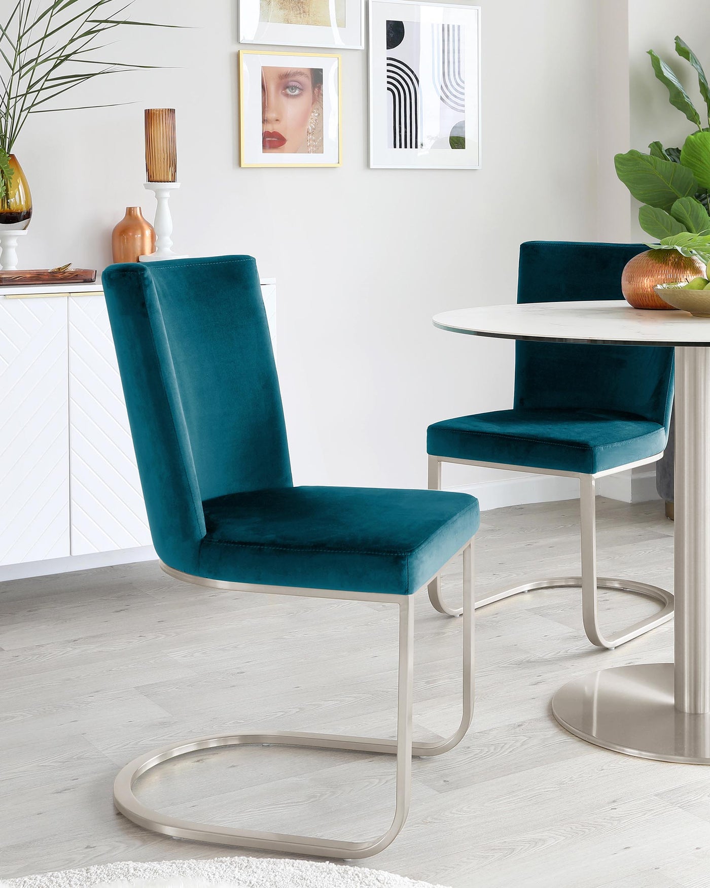 Elegant modern dining room featuring two sleek teal velvet chairs with a unique U-shaped metallic base, complementing a white round marble-top table with a cylindrical metallic base, set on a light wooden floor with a small white rug beneath.