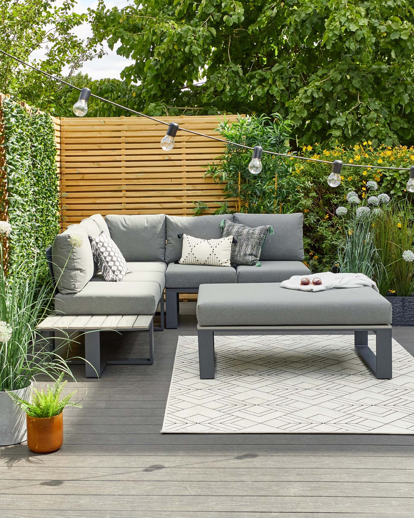 Modern outdoor furniture set featuring a dark grey sectional sofa with light grey cushions and various decorative pillows, accompanied by a matching dark grey rectangular ottoman serving as a coffee table. Set on a geometric patterned outdoor rug.
