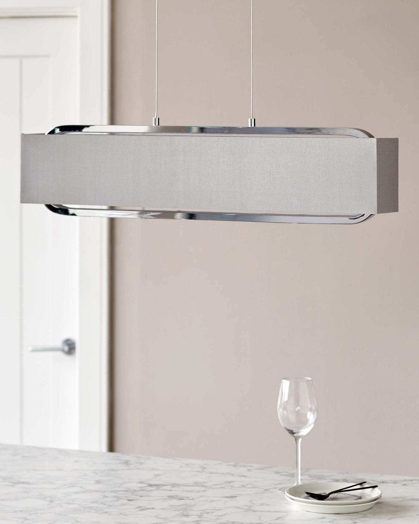 Elegant, contemporary pendant light with a sleek metallic and fabric finish, suspended above a white marble countertop with a wine glass and silverware set on a small plate, adding a touch of sophistication to a modern interior design.