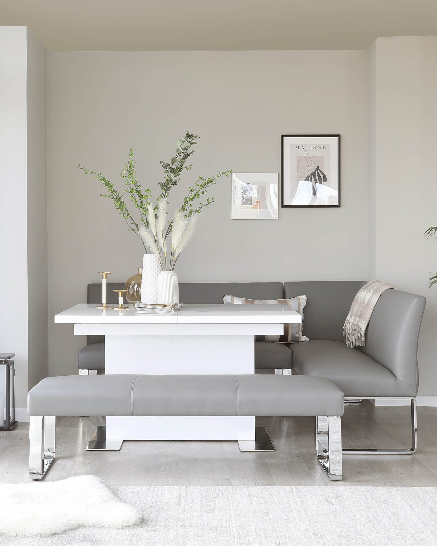 Modern minimalist dining room furniture set featuring a sleek white rectangular table with a broad base, paired with an elegant grey upholstered bench and complementing L-shaped corner bench, both accented with chrome-finished legs. A plush white area rug partially underlays the ensemble.