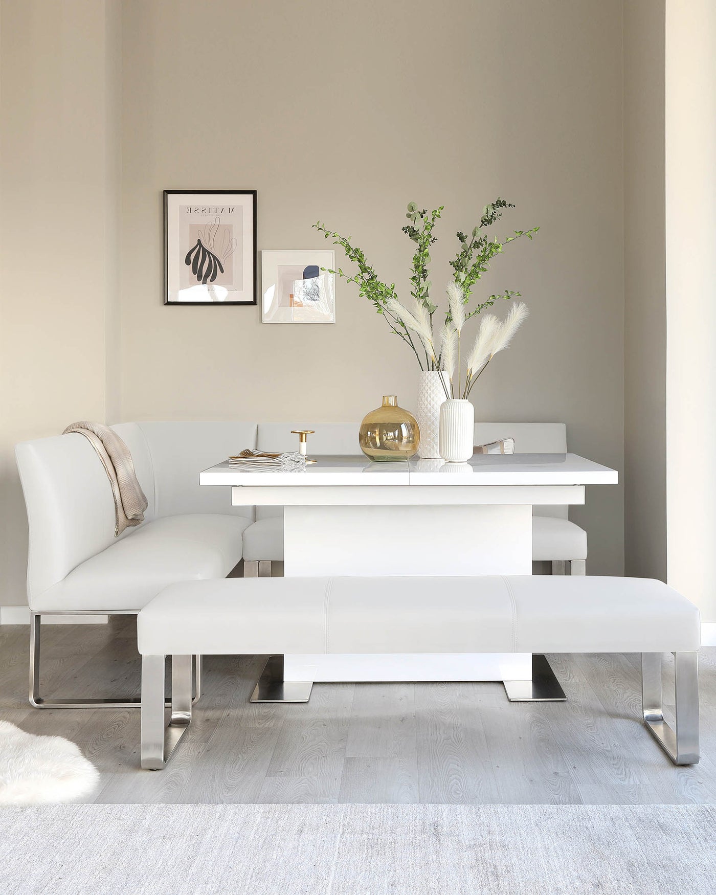A modern minimalist dining set featuring a white rectangular table with a glossy finish and sleek chrome legs. Accompanying the table is a white leather corner bench with clean lines and chrome legs, flanked by a matching white leather ottoman. The setting is complemented by a soft white rug beneath.