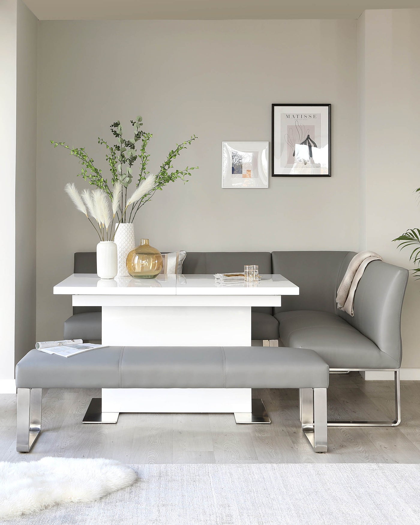 Modern minimalist furniture set in a neutral-coloured living space, featuring a sleek white rectangular dining table with a glossy finish, paired with a grey upholstered bench with chrome metal legs. The scene is accessorized with decorative elements, including greenery and art frames, creating an elegant and sophisticated ambiance.