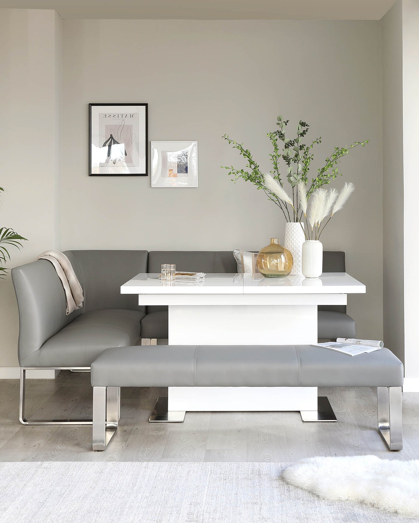 Modern dining area featuring a white, minimalistic dining table with a matching bench and grey upholstered L-shaped corner seat with metal accents on the legs.