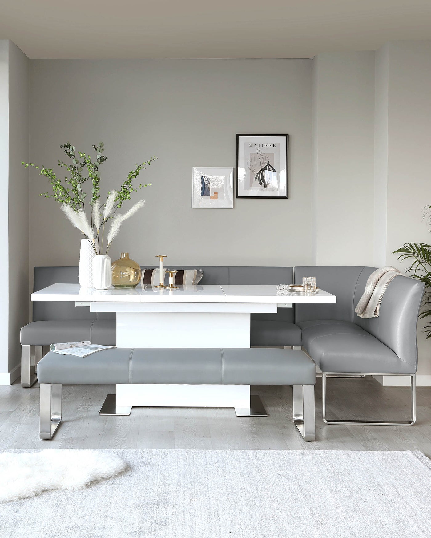 Modern dining area with a sleek white rectangular table featuring metallic accents, accompanied by a matching bench and a cantilevered grey upholstered chair with a stainless steel base. A plush white area rug lies underneath, complementing the contemporary aesthetic.