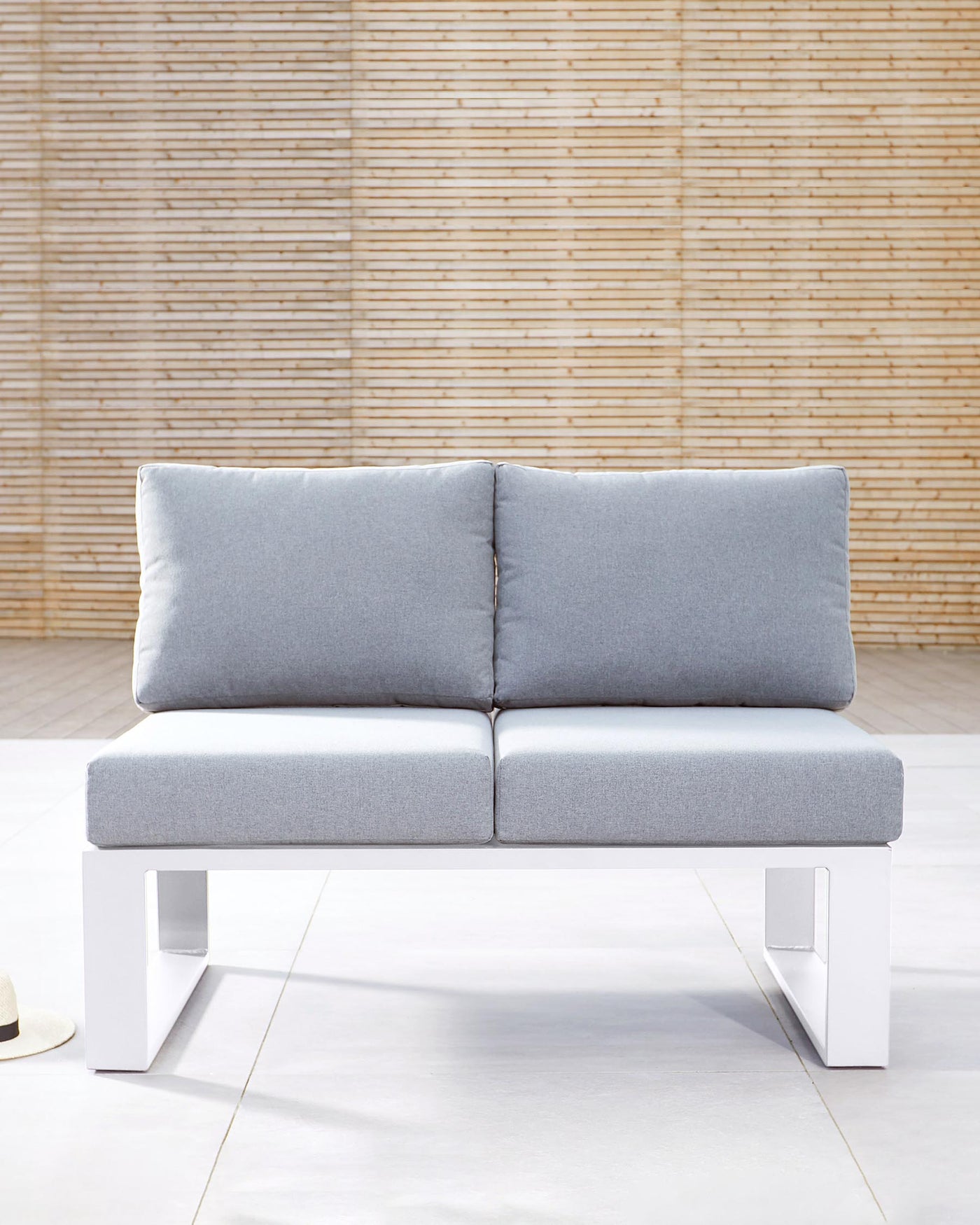 A modern minimalist two-seater sofa with a clean design, featuring a solid white base and light grey cushions. The base has a distinctive U-shaped silhouette that supports a pair of thick, comfortable seat cushions and two backrest cushions, all upholstered in a light grey fabric. The couch is showcased against a textured bamboo wall, suggesting a contemporary and natural ambiance.
