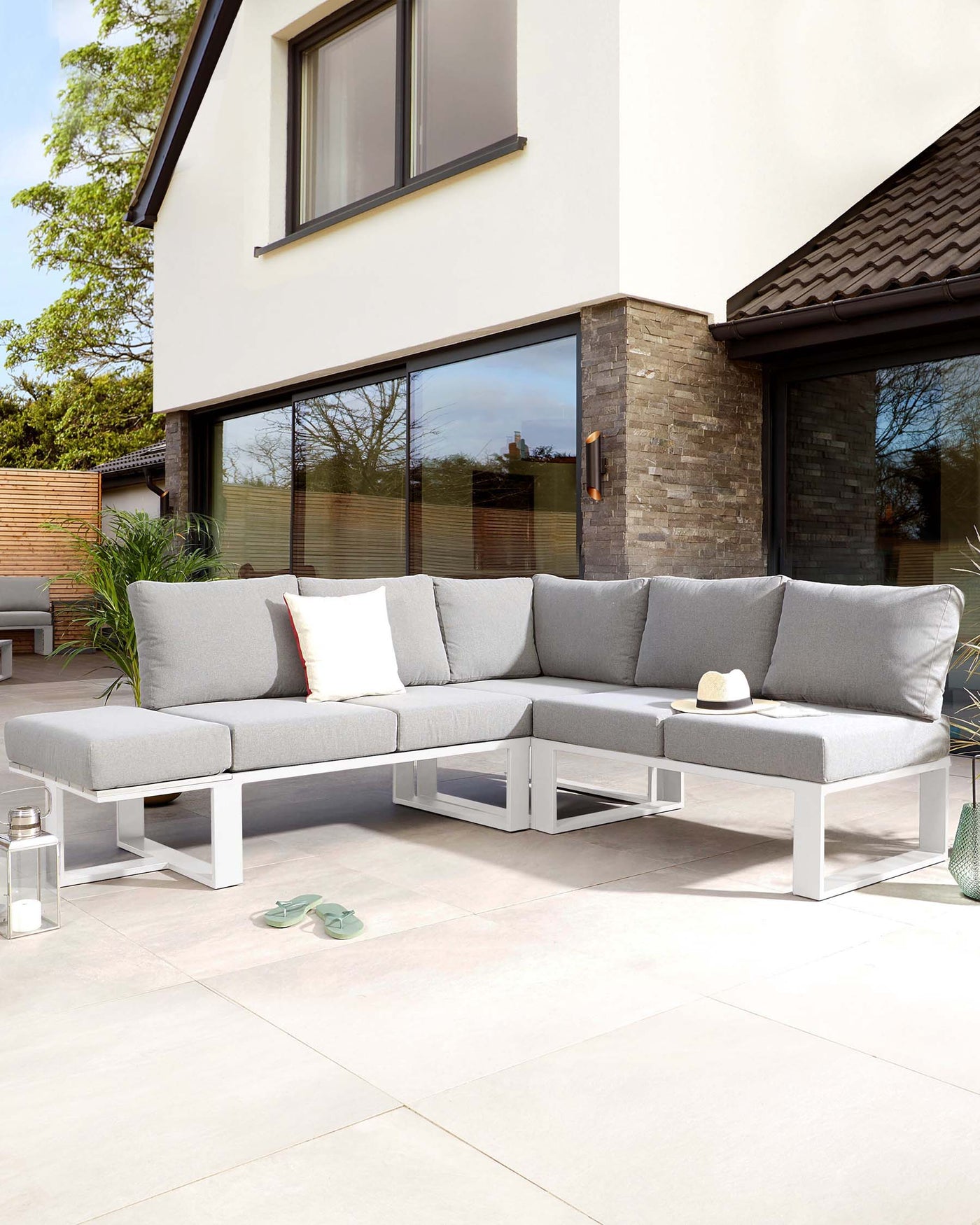 Modern outdoor sectional sofa with light grey cushions on a sleek white aluminium frame, complemented by a matching white low-profile coffee table. The ensemble is accessorized with assorted throw pillows in shades of grey and white, situated on a tiled patio.