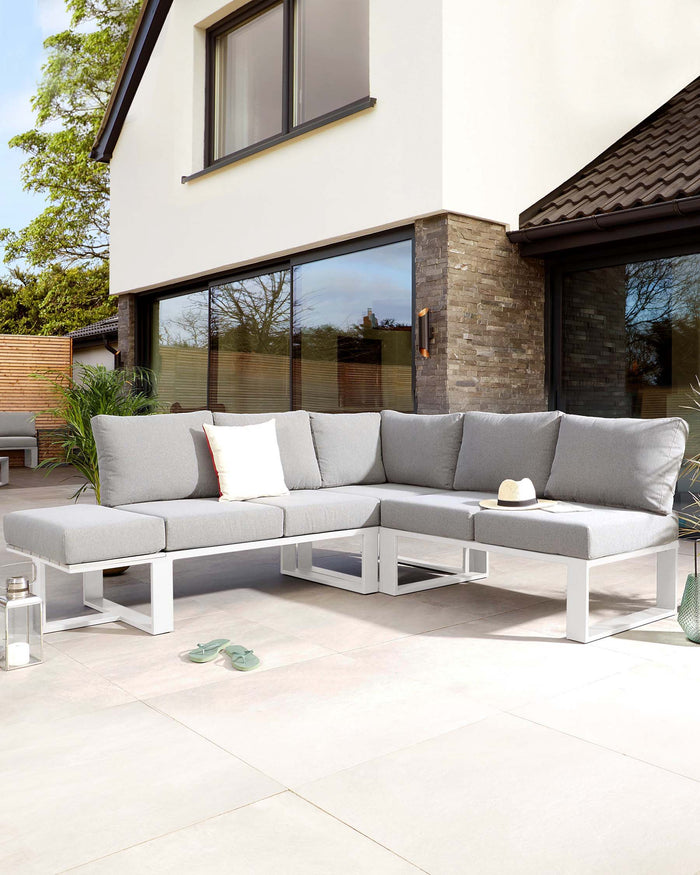Modern outdoor sectional sofa with white aluminium frames and grey cushions, complemented by white and red accent pillows, set on a patio. Two lantern-style candle holders sit on the ground beside the sectional.