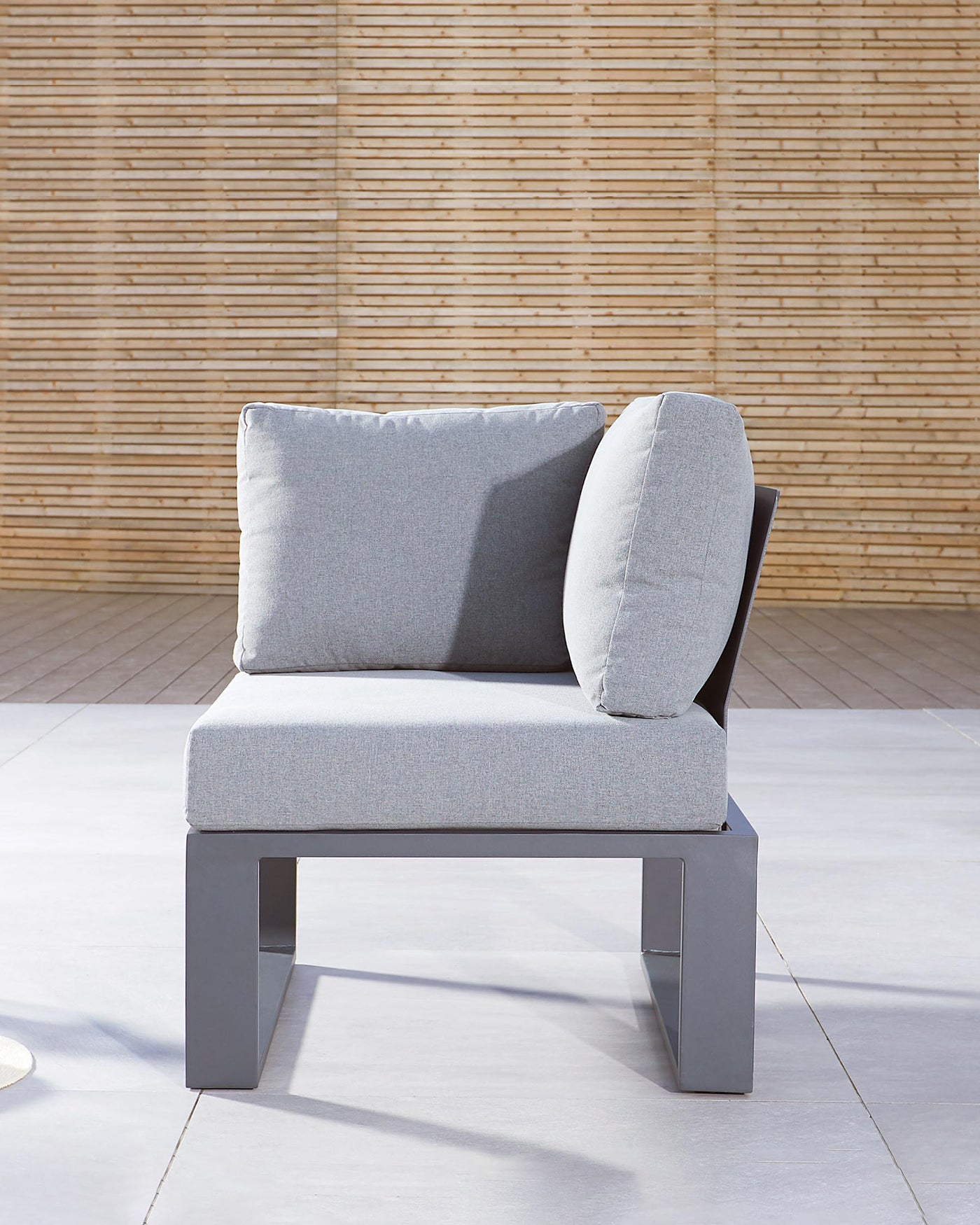 Modern single-seater sofa with a light grey fabric cushioned seat and backrest, complemented by a cylindrical side cushion, featuring a sleek metallic base and set against a warm bamboo backdrop.