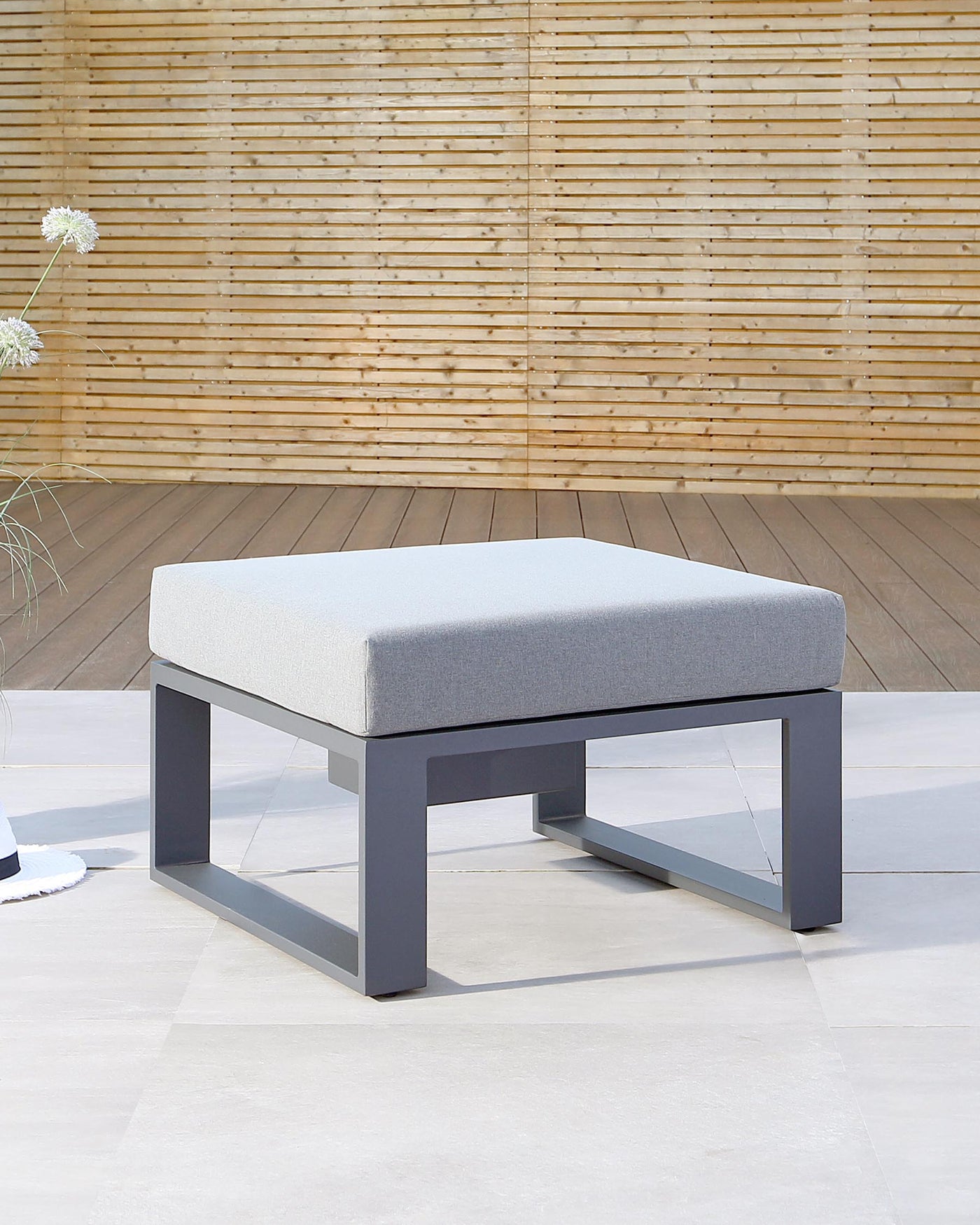 Modern outdoor ottoman with a grey cushion on a sleek, dark metal frame, displayed against a wooden slat backdrop and a tiled floor.