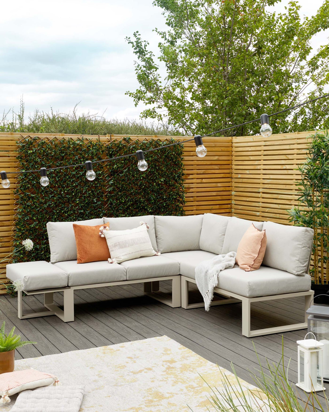 L-shaped modular outdoor sectional sofa with light grey cushions on a white aluminium frame, paired with decorative pillows in shades of orange and white with tassel accents, complemented by a cosy off-white throw blanket. A light grey and yellow patterned outdoor area rug rests underneath, while a white lantern sits nearby to complete the inviting patio arrangement.