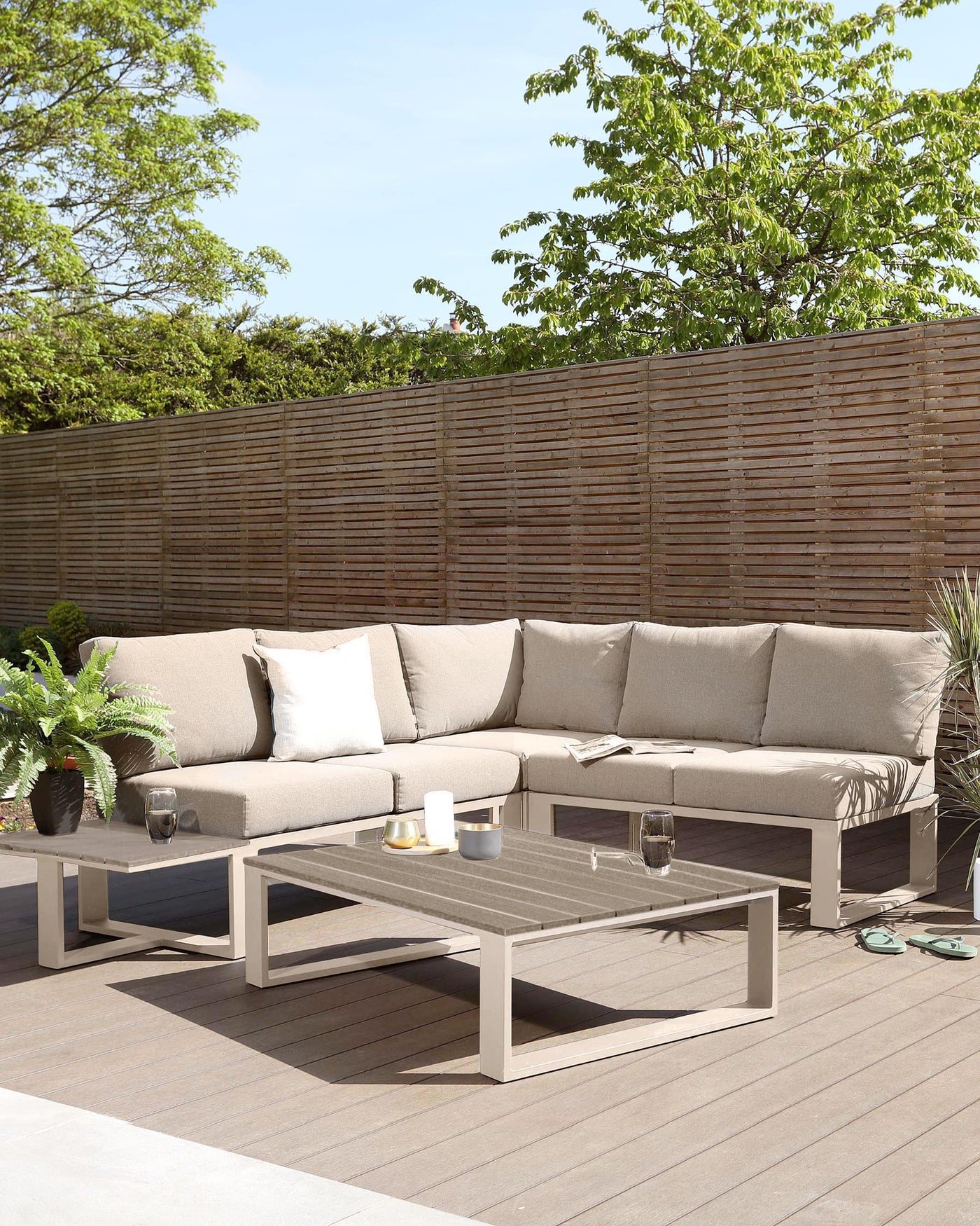 Outdoor patio furniture featuring a modular L-shaped sectional sofa with light taupe cushions and a matching low-profile rectangular coffee table with a slatted top, both with a minimalist metallic frame in light grey, set on a wooden deck.
