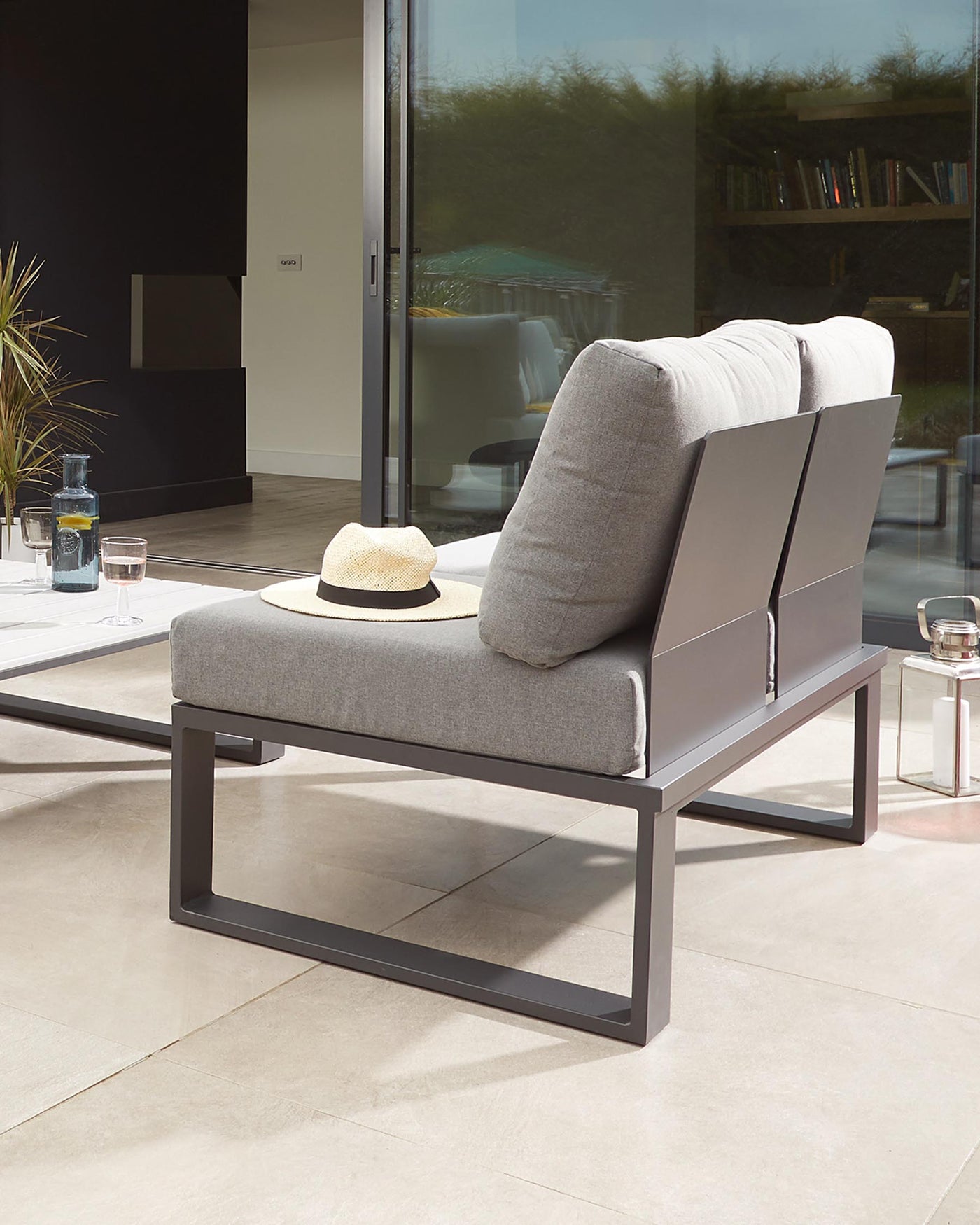 Modern outdoor chaise lounge with a minimalist metal frame in a dark finish and thick, plush cushions in a light grey fabric. The design features clean lines and a sleek, rectangular silhouette that complements a contemporary patio setting. A straw hat rests on the chaise, suggesting leisure and comfort.