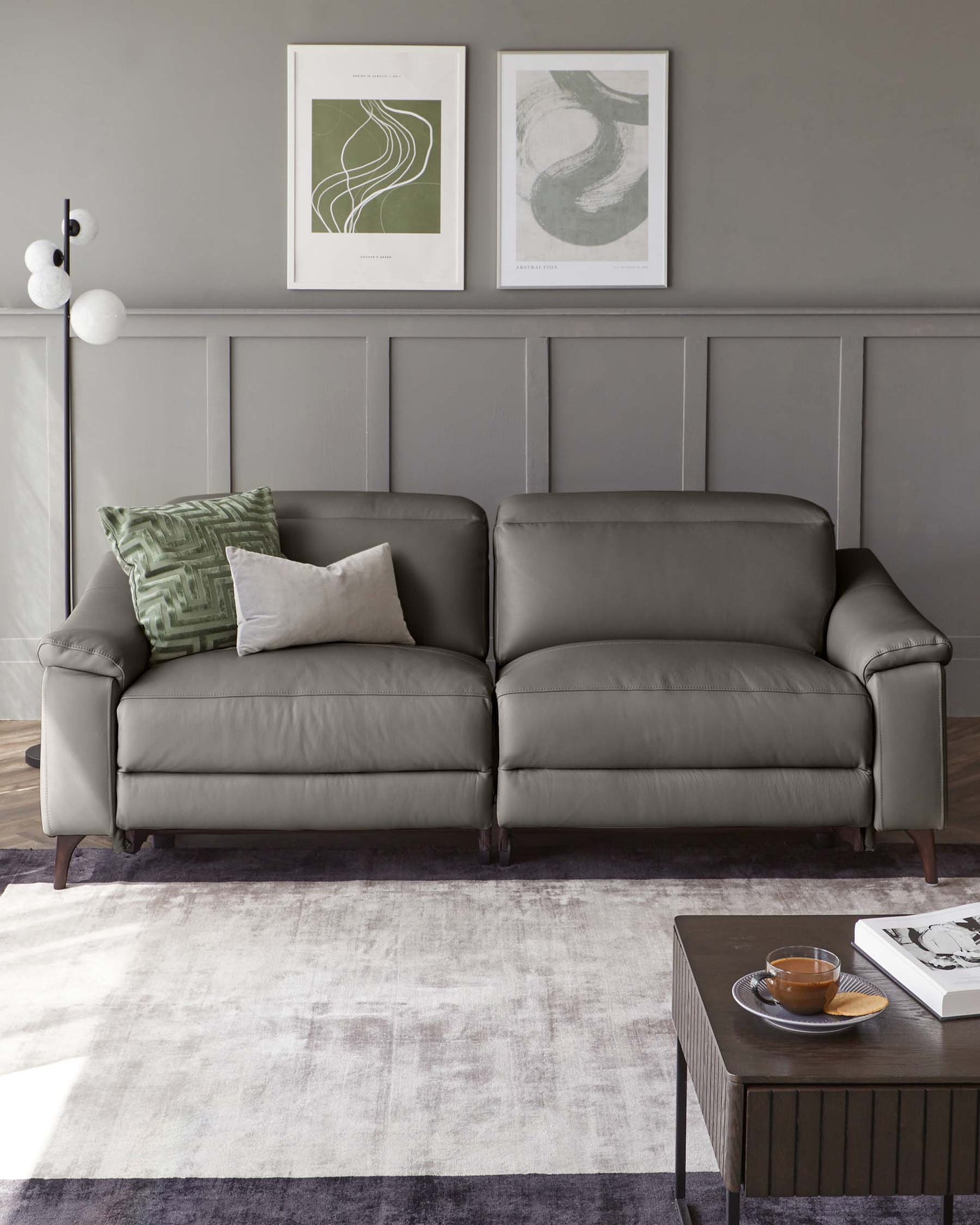 Modern grey upholstered sofa with plush cushions, accented by a light grey and white throw pillow, a green patterned pillow, and situated on a faded-grey area rug. Beside it is a contemporary dark wood coffee table with a glass teacup and saucer, and a book with a monochrome cover.