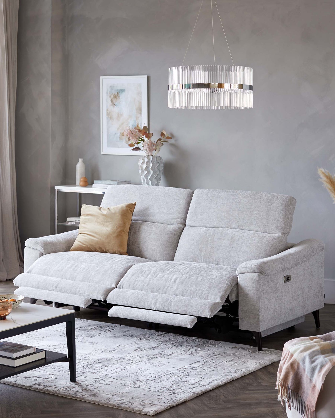 A contemporary light grey fabric reclining sofa with a tufted backrest and two extended footrests, complemented by a sleek black metal-framed coffee table with a white top, set upon a textured white and grey area rug.