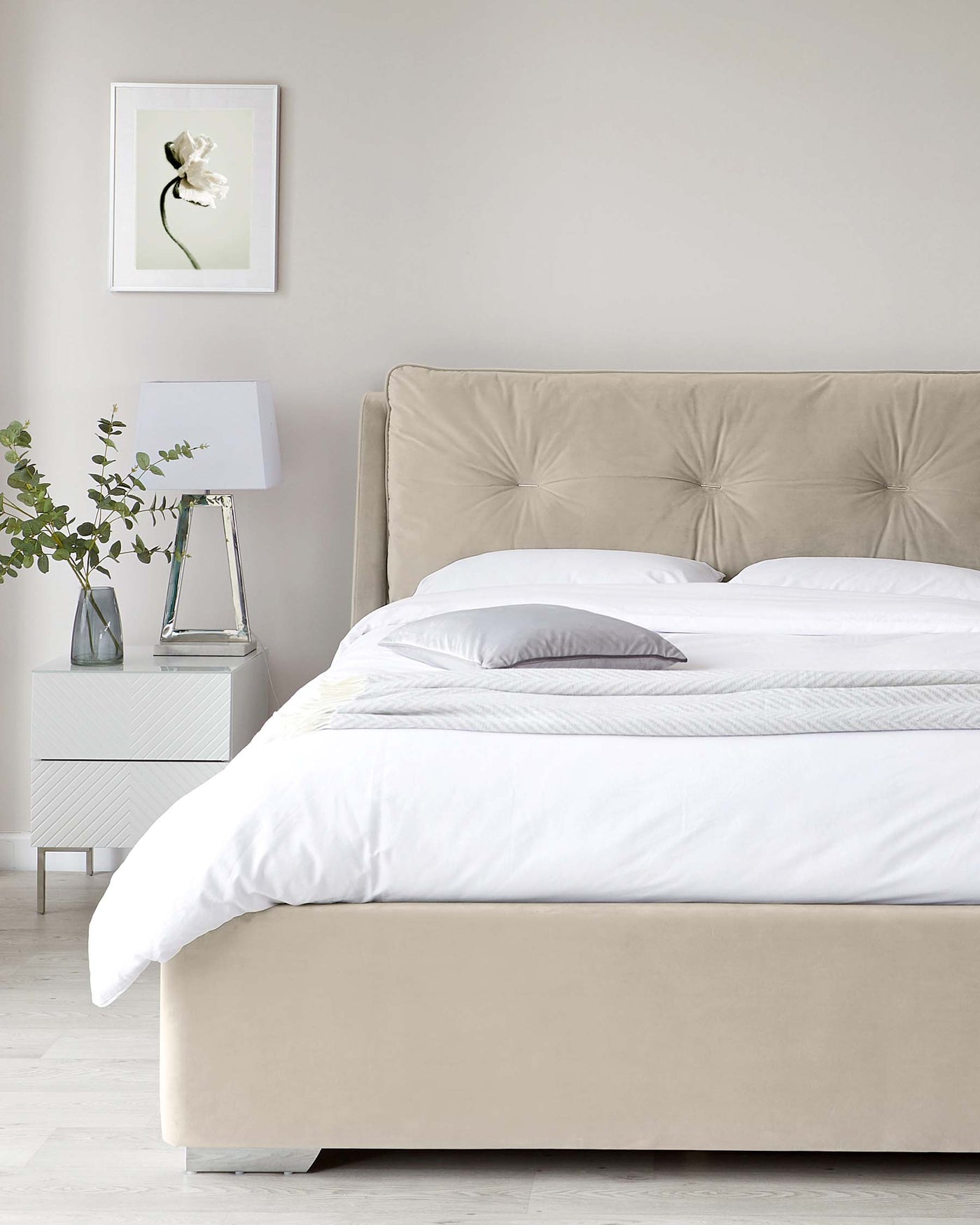 Elegant contemporary bedroom featuring a king-sized bed with an upholstered beige headboard showcasing button tufting, complemented by crisp white bedding. Beside the bed stands a sleek white nightstand topped with a modern clear glass lamp with a white lampshade.