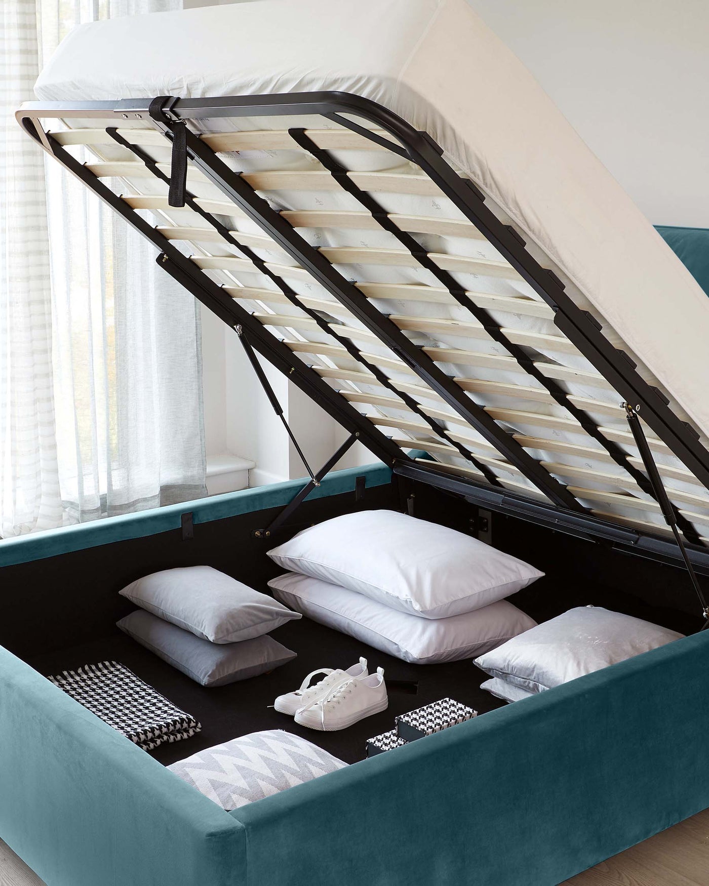 Teal upholstered storage bed with gas lift mechanism revealing spacious under-mattress storage containing pillows and personal items, set against a bright interior with sheer curtains.