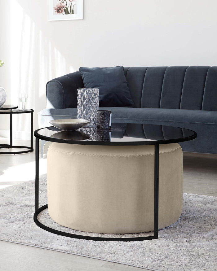 A contemporary living room setup featuring a dark grey velvet sofa with tufted backrest and a set of two round nesting coffee tables with black metal frames and smoked glass tops. Additionally, there is a round beige ottoman with a textured fabric finish placed on a light grey area rug. Decorative items, including vases and books, adorn the tables for a stylish ambiance.