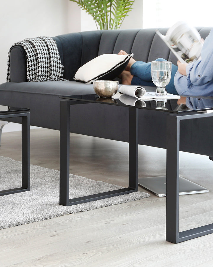 Modern living room interior featuring a sleek black upholstered sofa with a houndstooth-patterned throw and a contrasting white accent pillow. In front of the sofa is a contemporary black metal square coffee table with a glass top, displaying decorative items and a magazine. A plush grey area rug lies beneath the coffee table, complementing the light wood flooring.