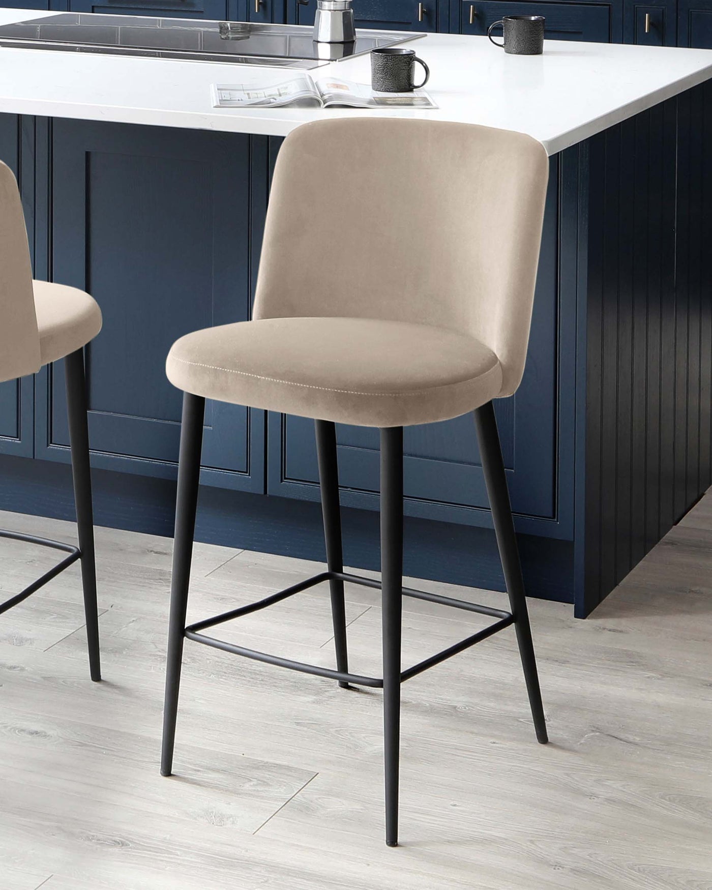 Two modern beige upholstered bar stools with sleek black metal legs, situated against a kitchen island with a white countertop and navy blue cabinetry.