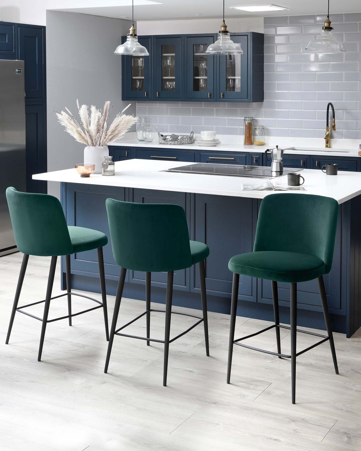 Three modern bar stools with plush, dark teal upholstery and sleek black metal legs, positioned at a white kitchen island.