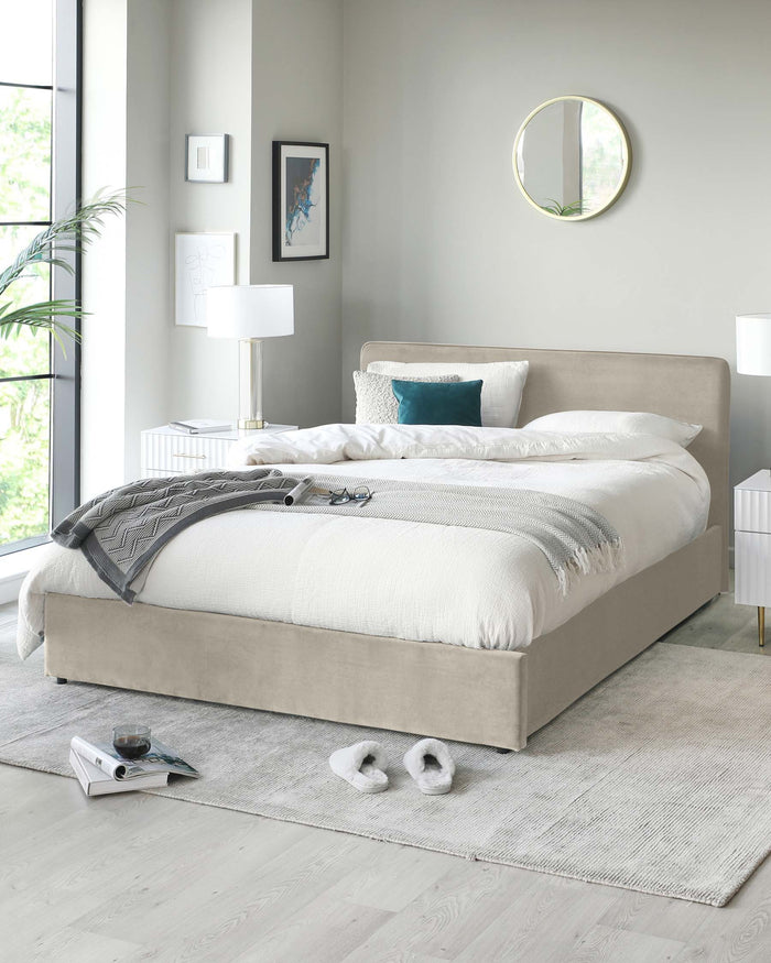 A contemporary bedroom featuring a beige upholstered platform bed with a gently curved headboard. Matching white bedside tables flank the bed, each adorned with a chic white lamp with a gold base. A round gold-framed mirror hangs on the wall, reflecting natural light from a window. The setting is grounded by a soft grey area rug, which complements the light hardwood floor.