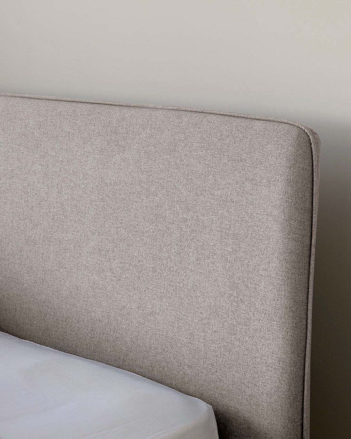 A close-up view of a contemporary upholstered headboard with a neutral heathered fabric finish, featuring a slightly curved top edge and clean, simple lines, suggesting a minimalist and modern aesthetic. The bedding or mattress is partially visible with a clean, plain white cover.