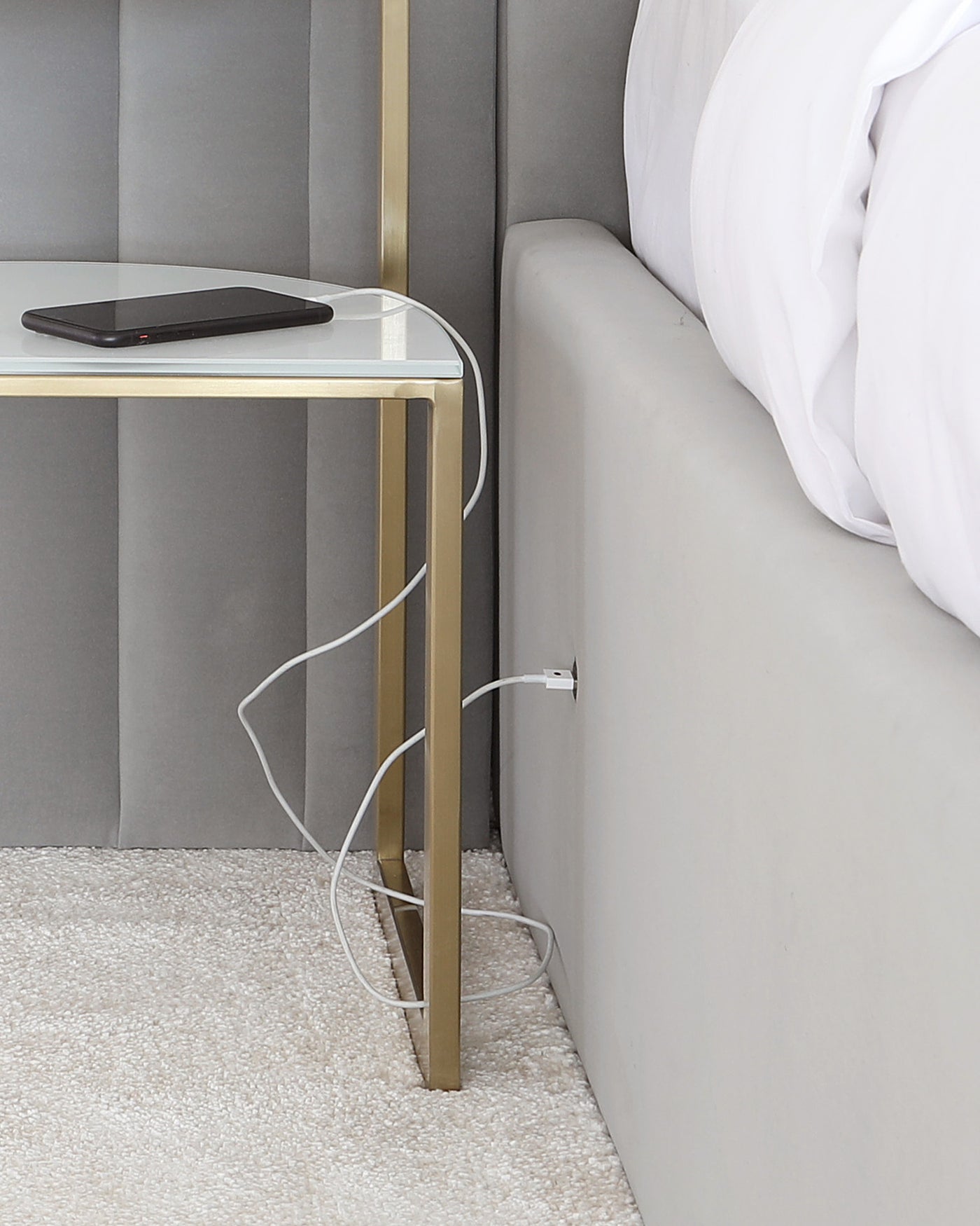 Elegant modern bedside table with a clear glass top and minimalist gold-tone metal frame, alongside a plush grey upholstered bed with crisp white bedding.