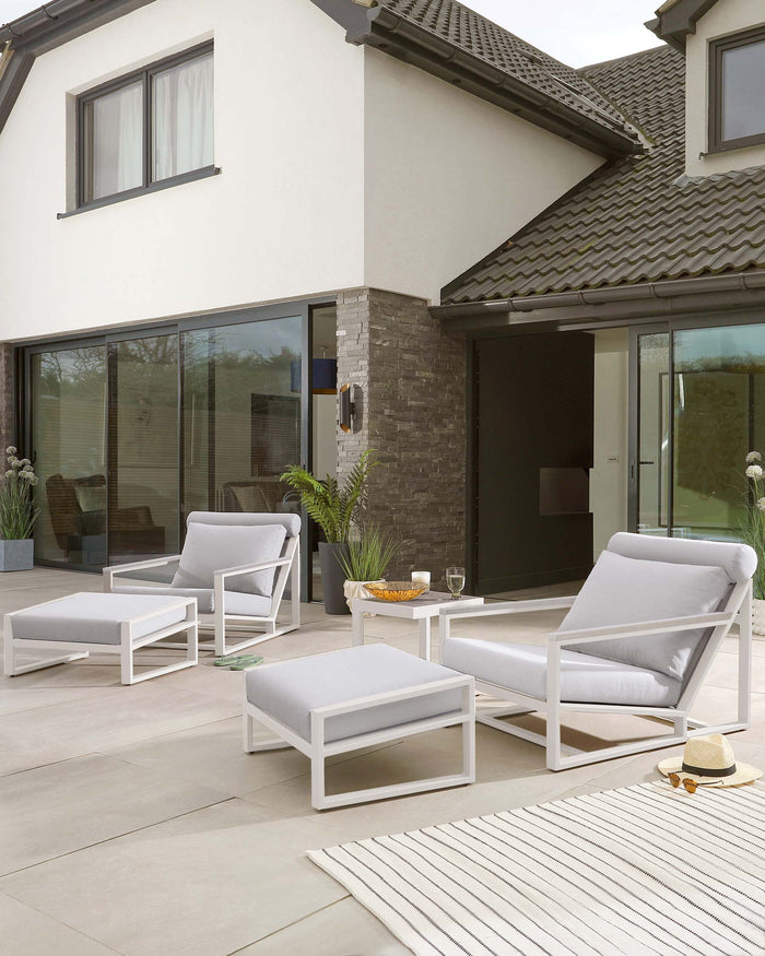 A modern outdoor furniture set consisting of two white-framed armchairs with grey cushions, complemented by a matching white-framed ottoman and a low white-framed coffee table with a clear glass top, arranged on a patio.