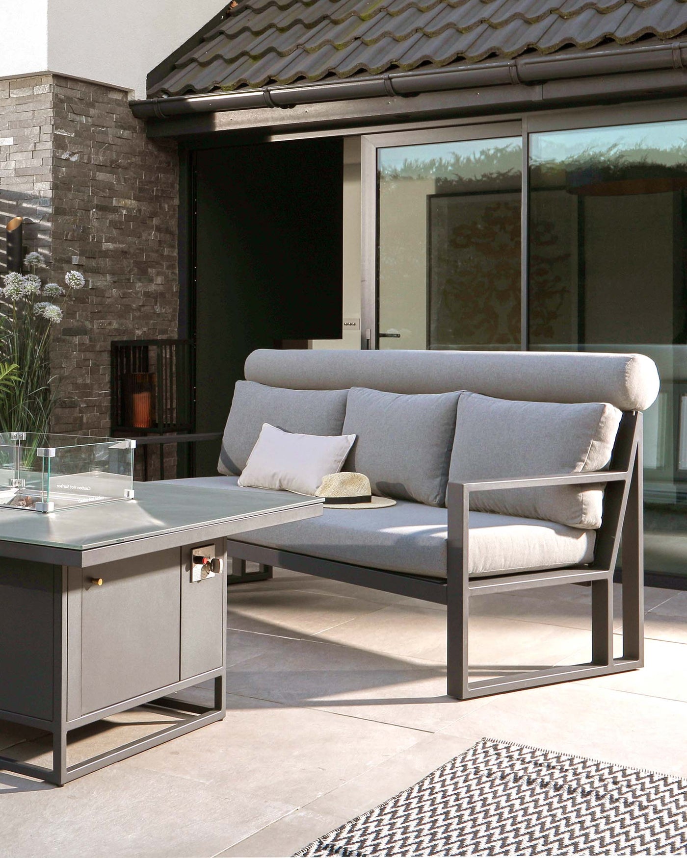 Contemporary outdoor furniture set featuring a sleek grey two-seater sofa with plush cushions and a matching grey rectangular coffee table with a glass top and built-in metal shelf. A simple white pillow and a wicker hat rest on the sofa, adding a touch of comfort and style. The set suggests sophisticated design suitable for modern patio or garden spaces.