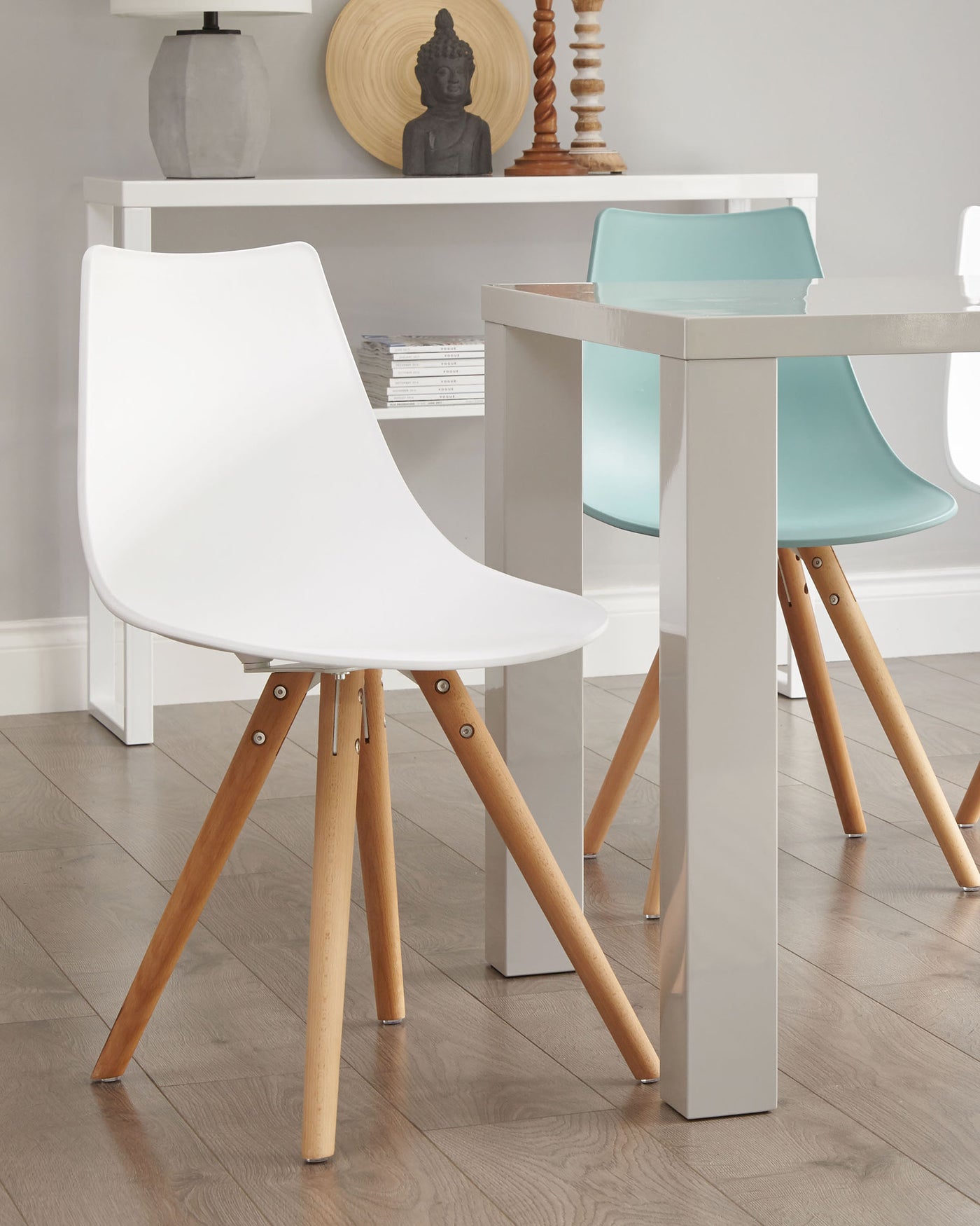 Modern dining room furniture featuring a white square table with sleek grey legs, paired with two minimalist chairs – one white and one turquoise – with smooth plastic seats and angled wooden legs connected by metal brackets. The setting displays a clean, contemporary aesthetic.