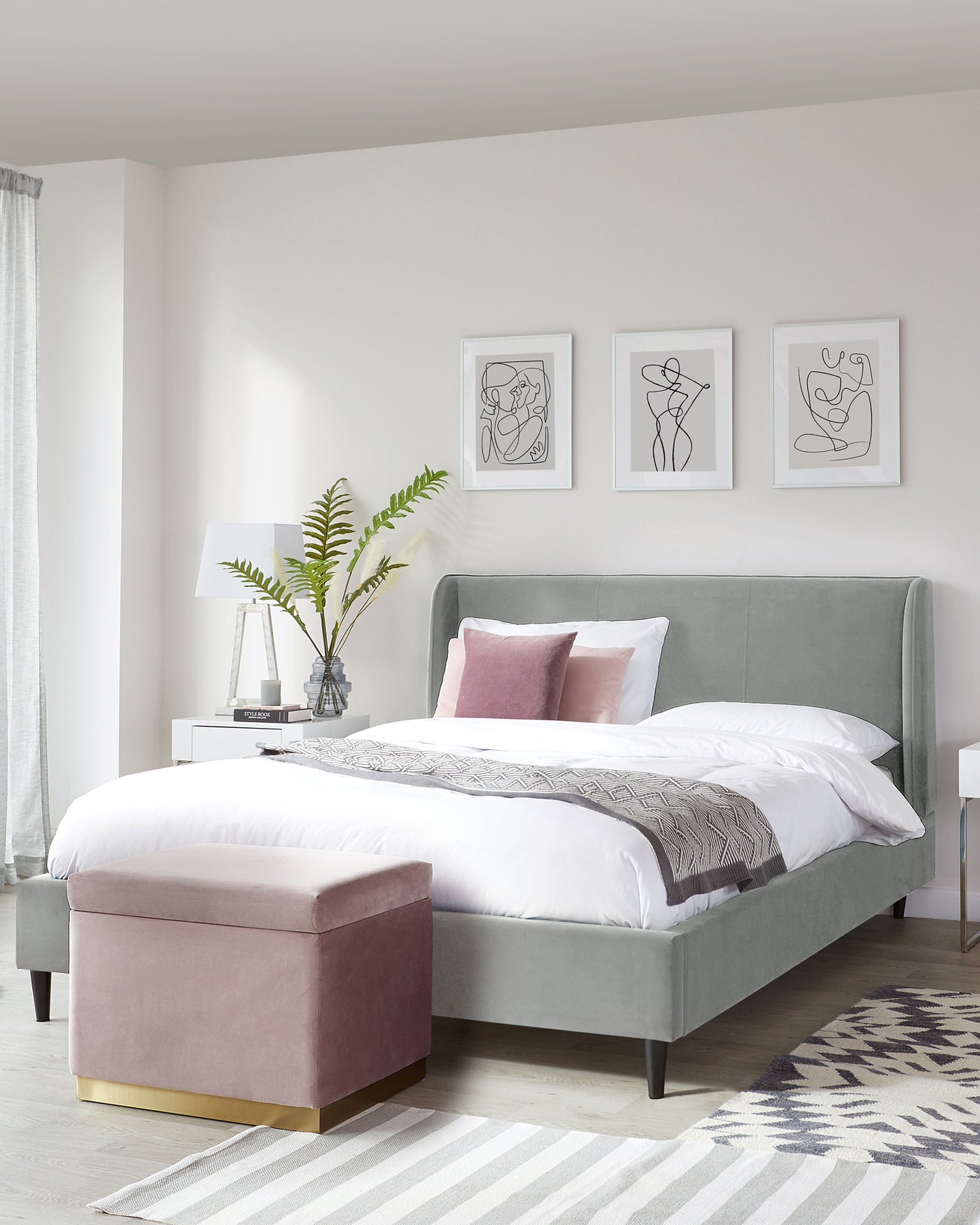 A contemporary styled bedroom featuring a grey upholstered bed with a high headboard, accompanied by a plush, dusty rose-pink storage ottoman with a gold metal base. The bed is dressed in white and patterned bedding, and a modern white nightstand with a table lamp and decorative elements completes the scene. The floor is adorned with a geometric patterned area rug with a complementing colour scheme, set against the backdrop of a neutral-toned room with framed abstract artwork on the wall.