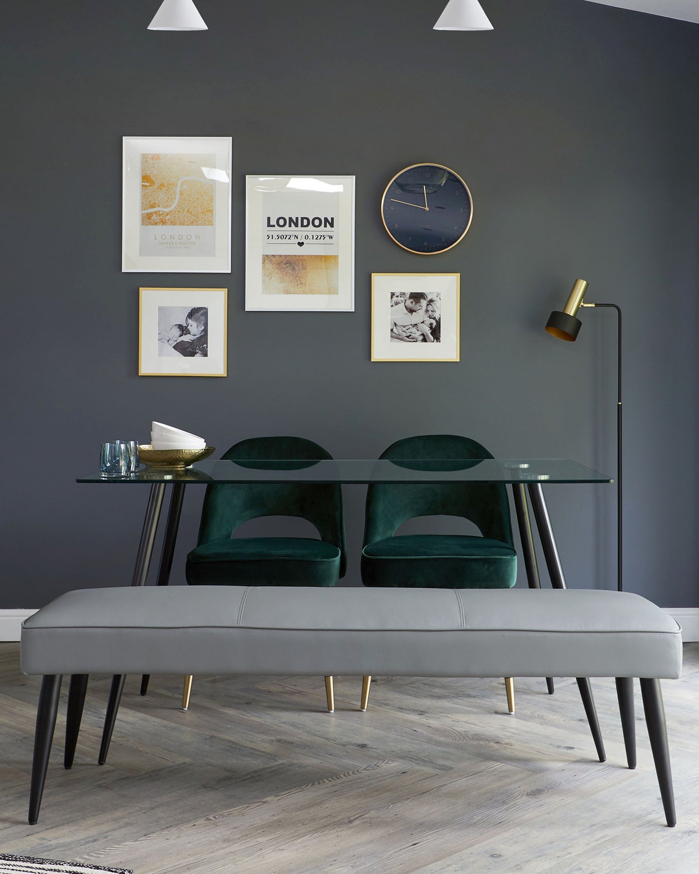 A modern dining set featuring a rectangular glass-topped table with black tapered legs, accompanied by four plush velvet green chairs. A sleek grey upholstered bench with black legs and gold feet accents sits to the forefront.