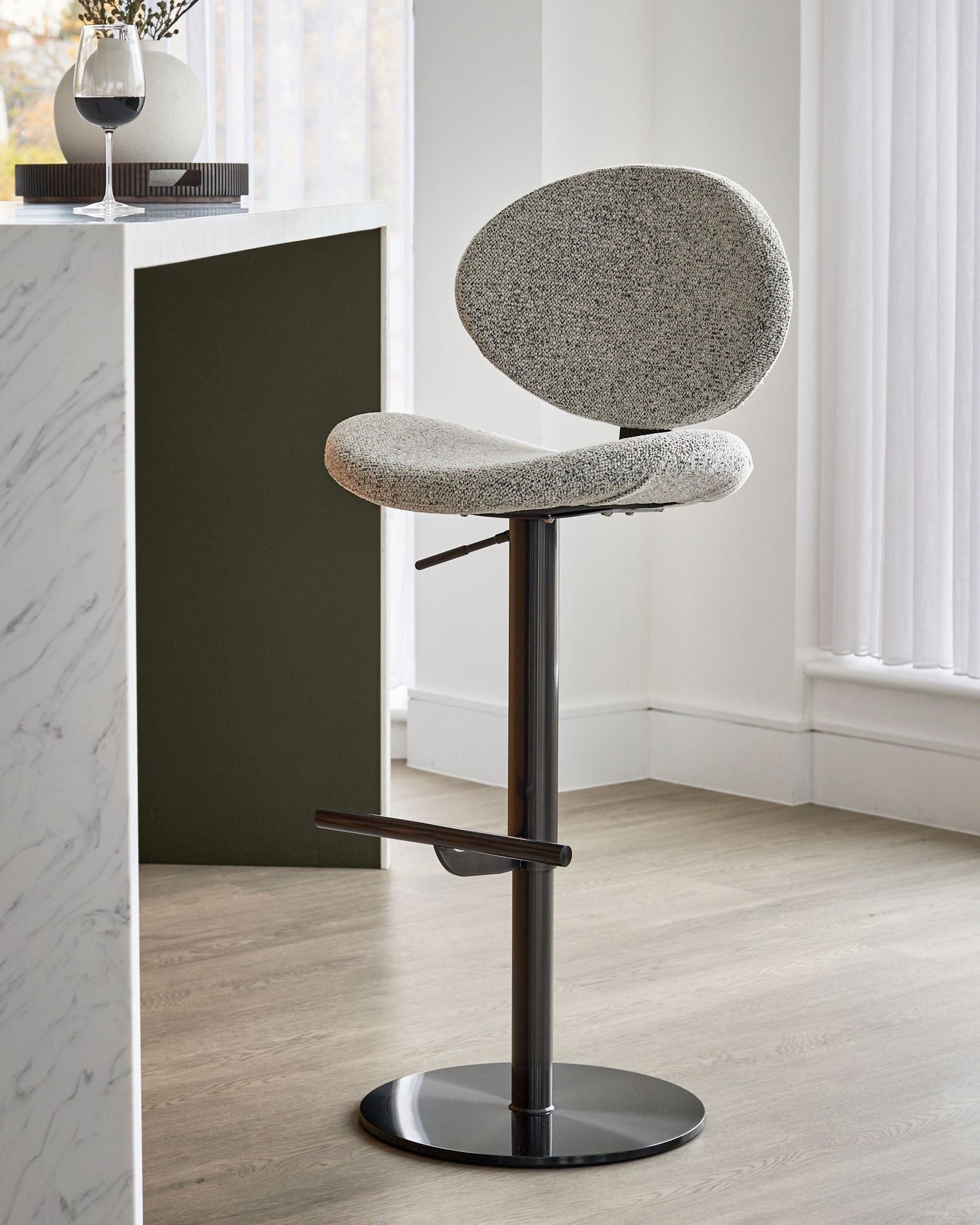 Lilah grey fabric bar stool with round backrest, curved seat, black metal base and footrest, adjustable height.