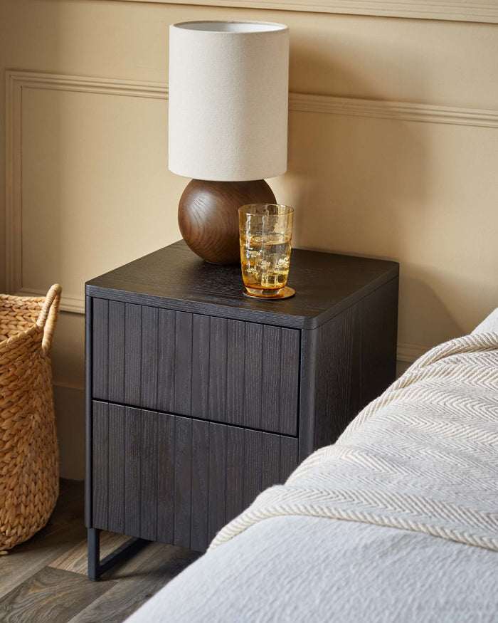 A dark wood bedside table with a lamp, glass of water, and a woven basket next to a bed.