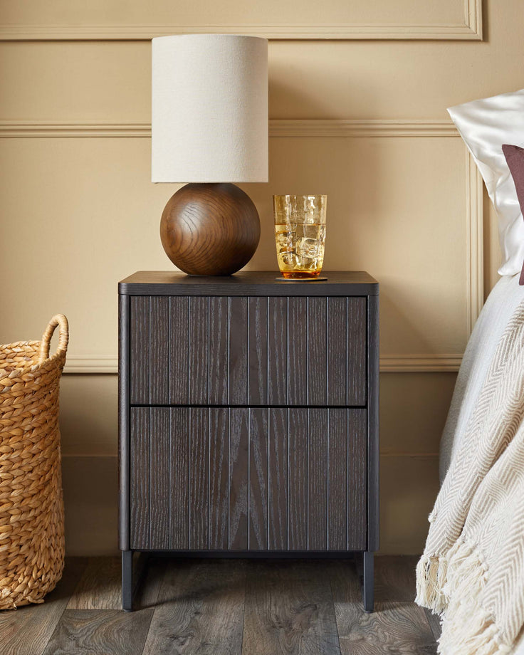 A dark wood bedside table with a lamp, glass and woven basket on a wood floor in a bedroom setting.