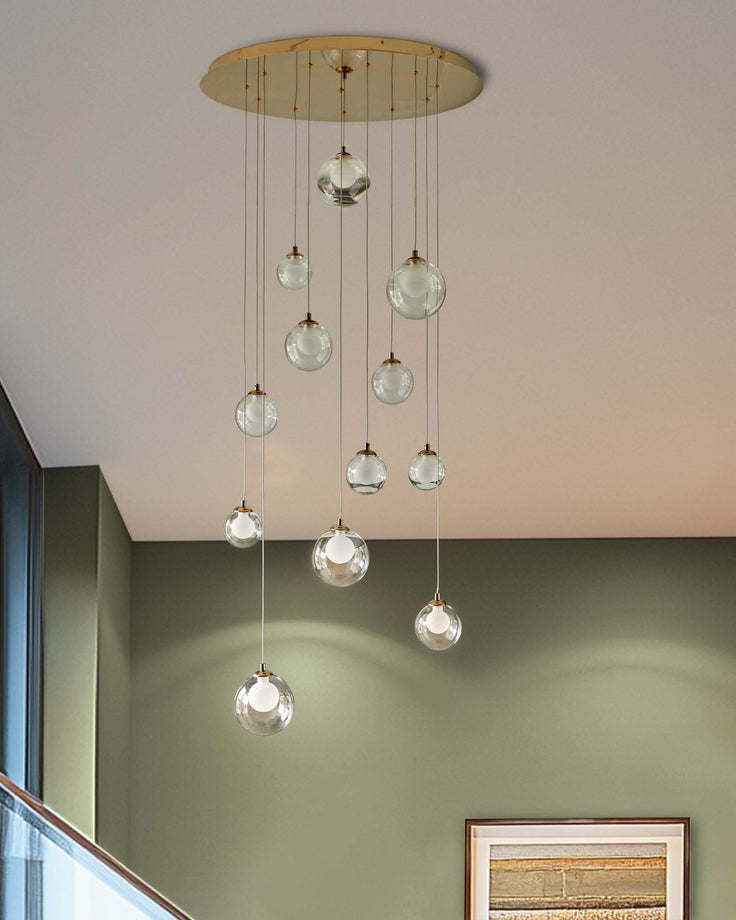 A modern cascading chandelier with glass globe lights, hanging from a gold ceiling plate. The backdrop includes a muted olive-green wall.
