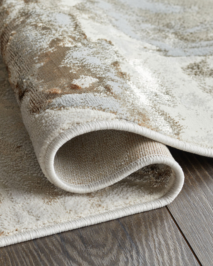 Beige and brown patterned rug, rolled at the edge to show underside, laying on dark wood floor.