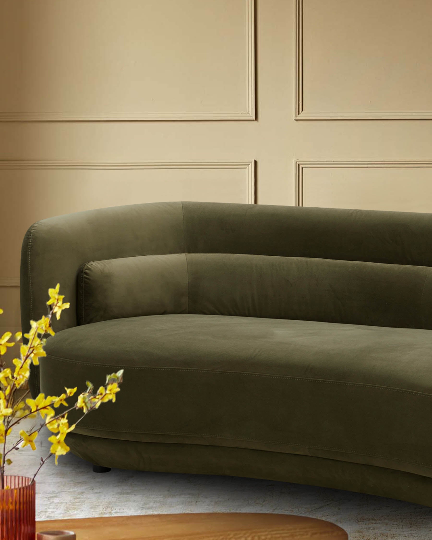 Olive green velvet sofa with curved back against a wall. Vases of yellow flowers decorate a table in the foreground.
