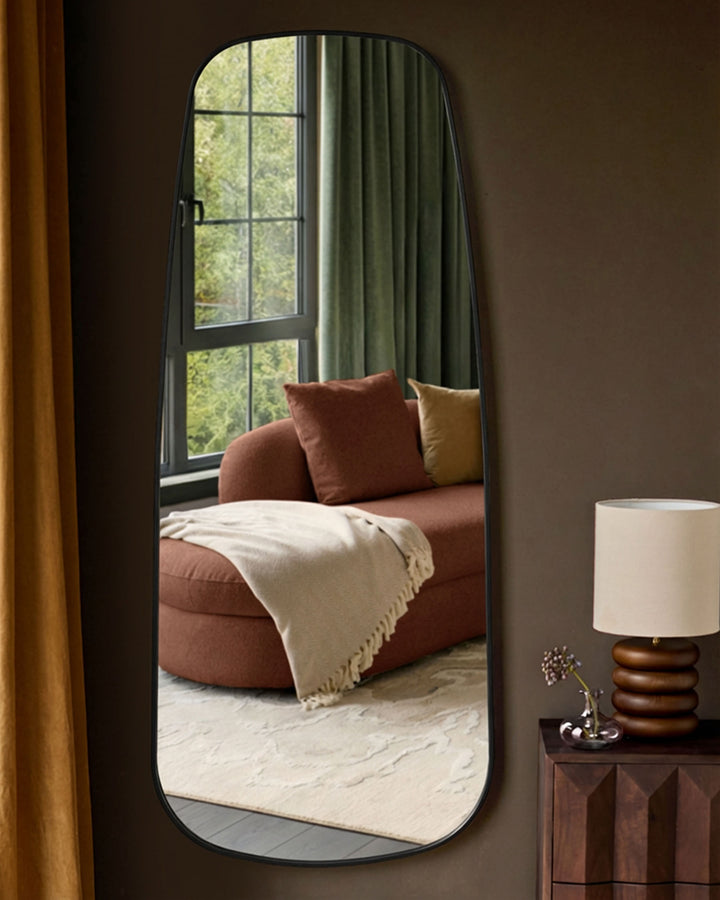 Black rounded rectangular full-length leaner mirror reflecting a rust-colored sofa and window with green curtains.