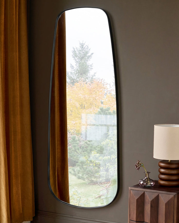 Black rounded rectangular full-length leaner mirror displayed against a Grey wall, reflecting outdoor garden view.