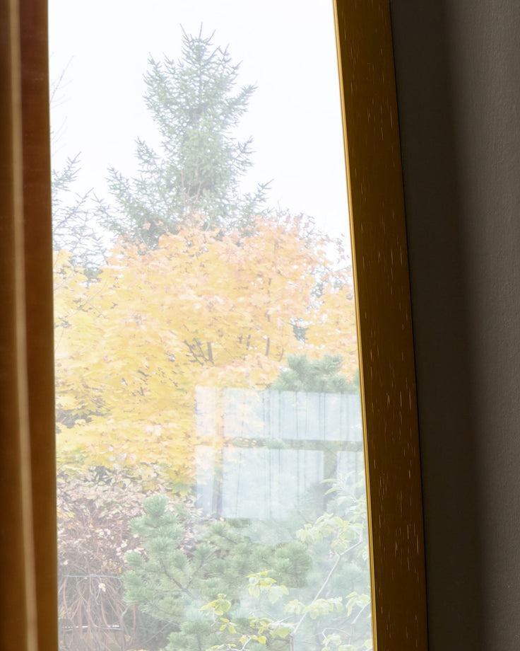 Gold full-length leaner mirror reflecting autumn garden view with trees and fence.