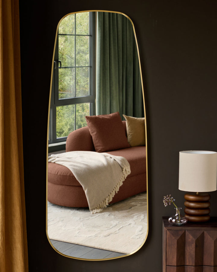 Gold-framed full-length leaner mirror with rounded edges, leaning against dark wall in modern bedroom setting.