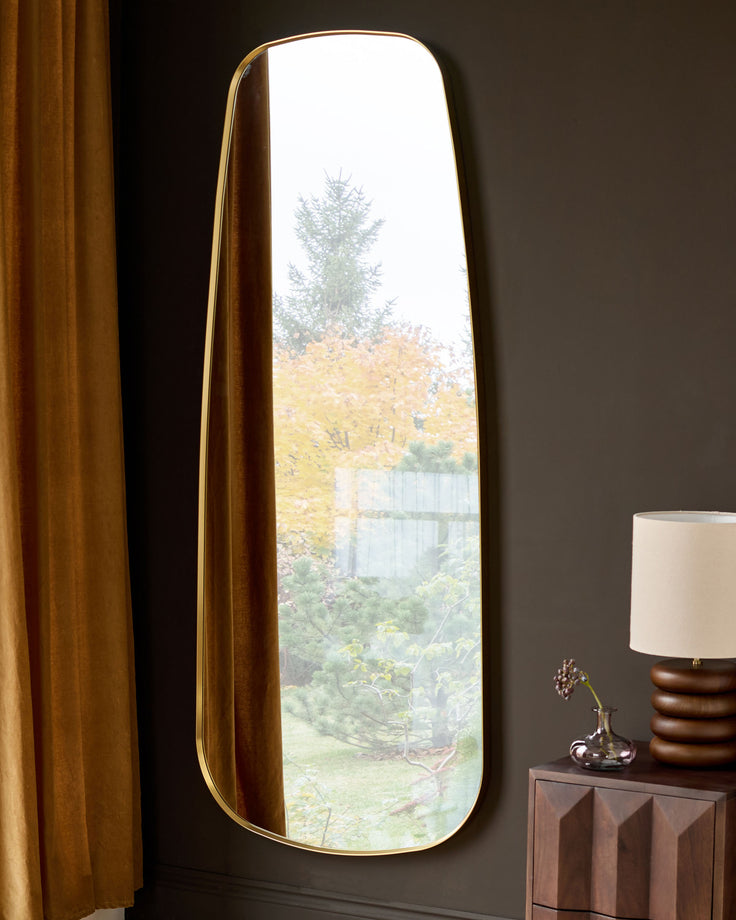 Gold-framed arched full-length leaner mirror against dark wall, reflecting garden view and interior decor.