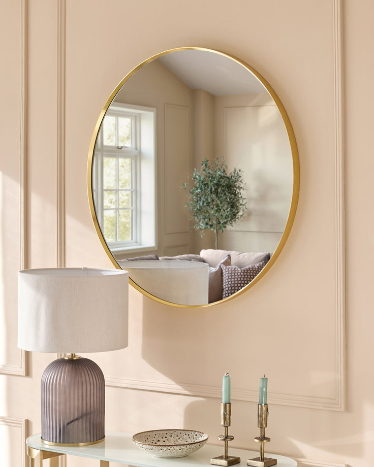 Large round gold-framed wall mirror reflecting a bright room with window, styled with lamp, candles, and decorative bowl.