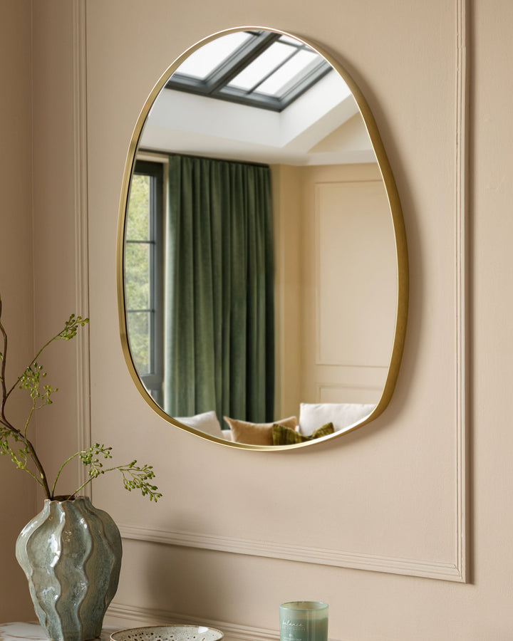 Gold oval wall mirror with rounded pebble shape, displayed above a console table with decorative vase and candle.