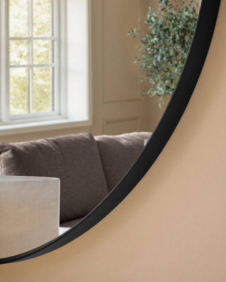Large round black-framed wall mirror reflecting a living room with Grey sofa, window, and potted plant.