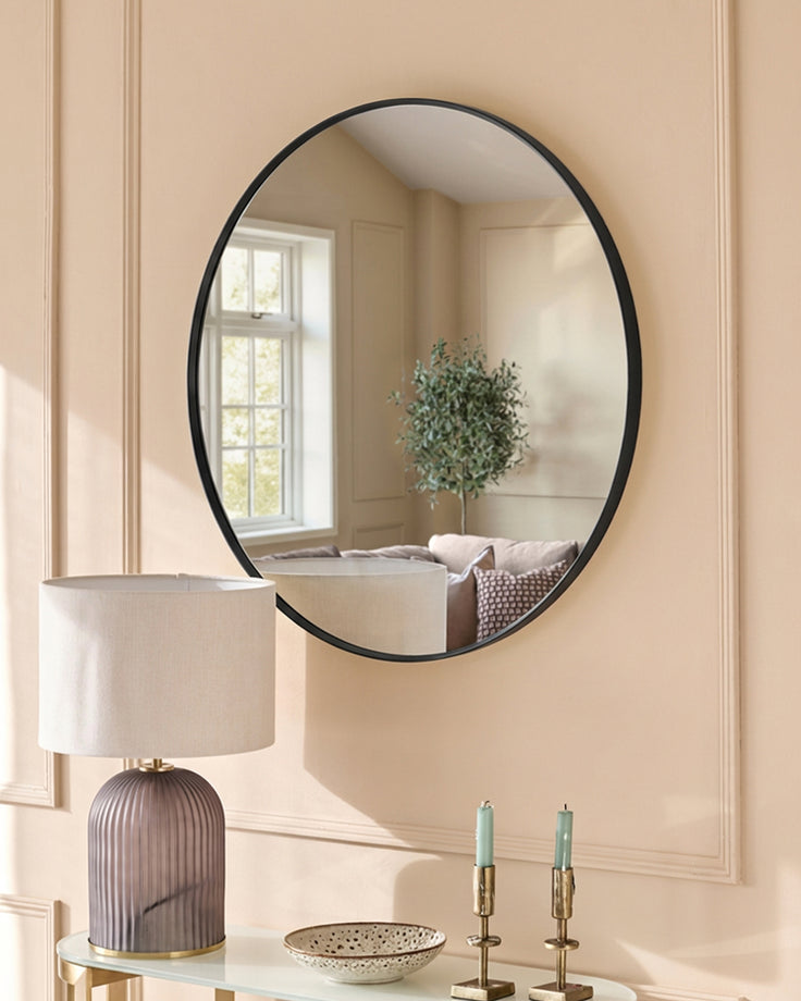 Large oval black-framed wall mirror displayed above a console table with lamp, candles, and decorative bowl in a neutral room.