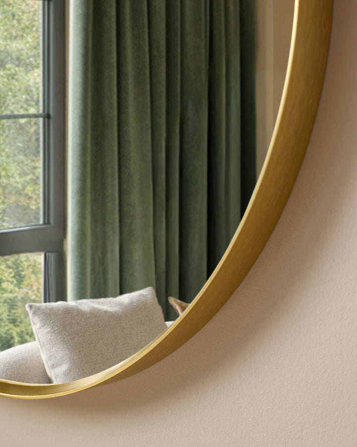 Gold curved wall mirror with elegant arched frame, displayed above a beige upholstered chair near windows with green curtains.