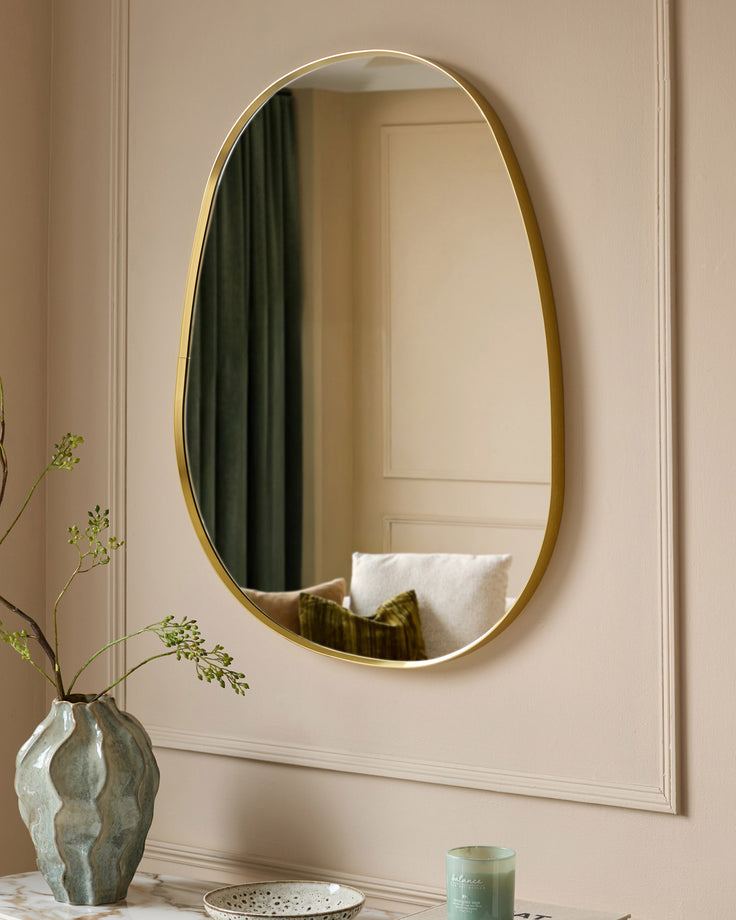 Gold oval wall mirror with curved frame, displayed above a table with decorative vase and accessories.
