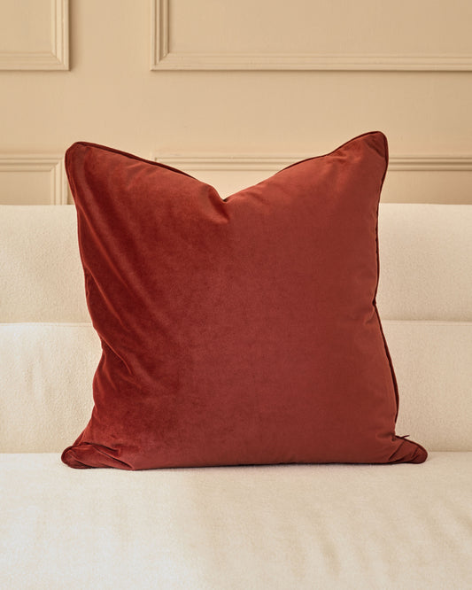 Large toffee red velvet cushion with piping detail, displayed on a cream upholstered bed against a paneled wall.